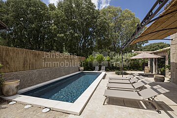 A large swimming pool is surrounded by lawn chairs and umbrellas and a sign that says janssens immobilier