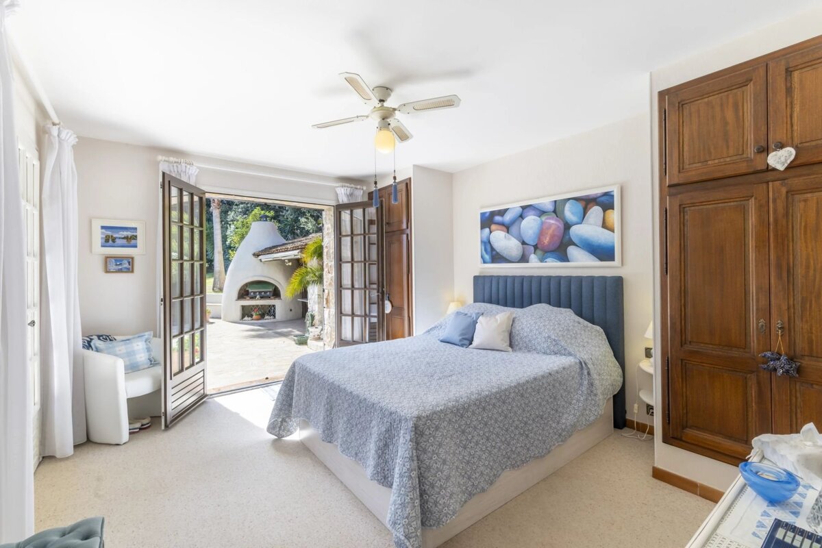 Bright bedroom with a blue bed, ceiling fan, and pebble artwork. French doors open to a sunny patio featuring an outdoor oven/fireplace.