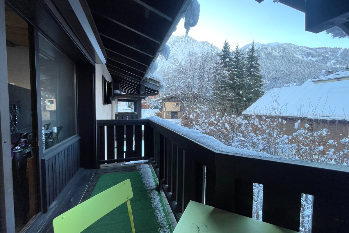 A balcony with a neon green chair and snow on its railing overlooks a picturesque, snow-covered mountain village with pine trees under a clear winter sky.
