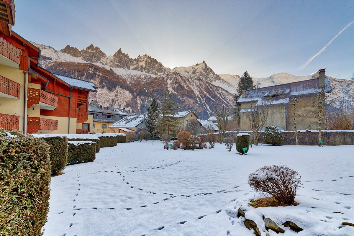 A snowy courtyard with buildings, footprints, and dramatic snow-capped mountains under a clear sky, likely a winter morning.