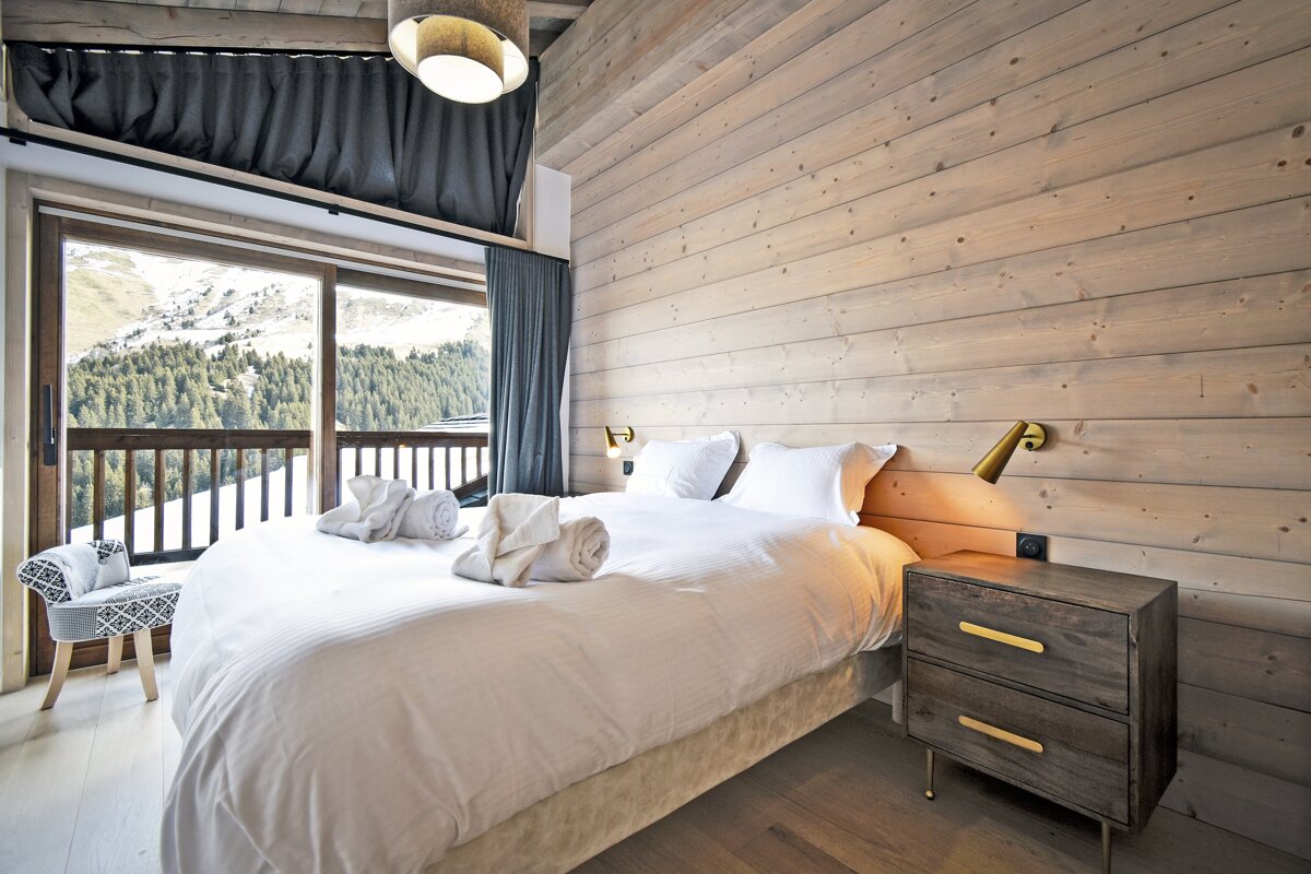 A cozy chalet bedroom with wood-paneled walls, a large bed, and a sliding glass door revealing a snowy mountain view.