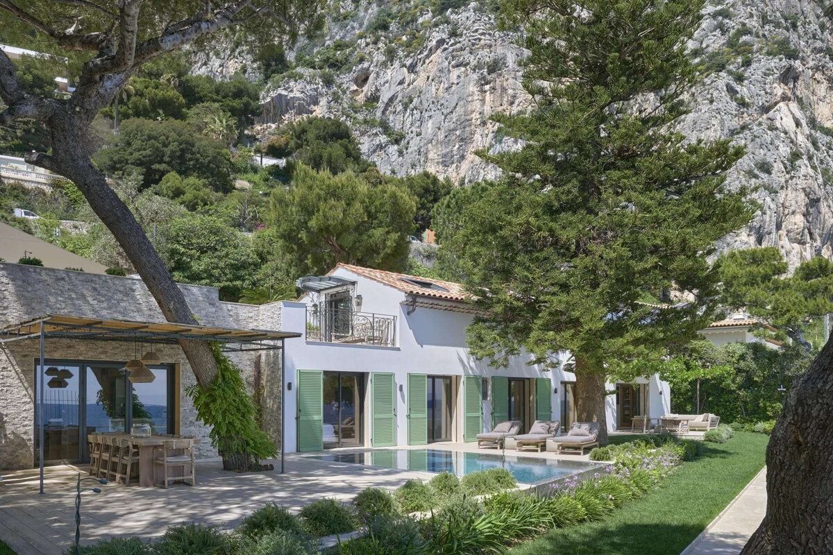 A white villa with a pool, lush garden, and outdoor dining area is nestled against a dramatic backdrop of rocky mountains and green foliage.