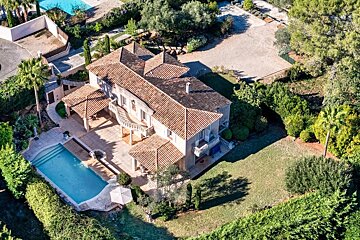 Aerial view of a grand house with a terracotta roof, a large swimming pool, and sprawling green grounds surrounded by trees.