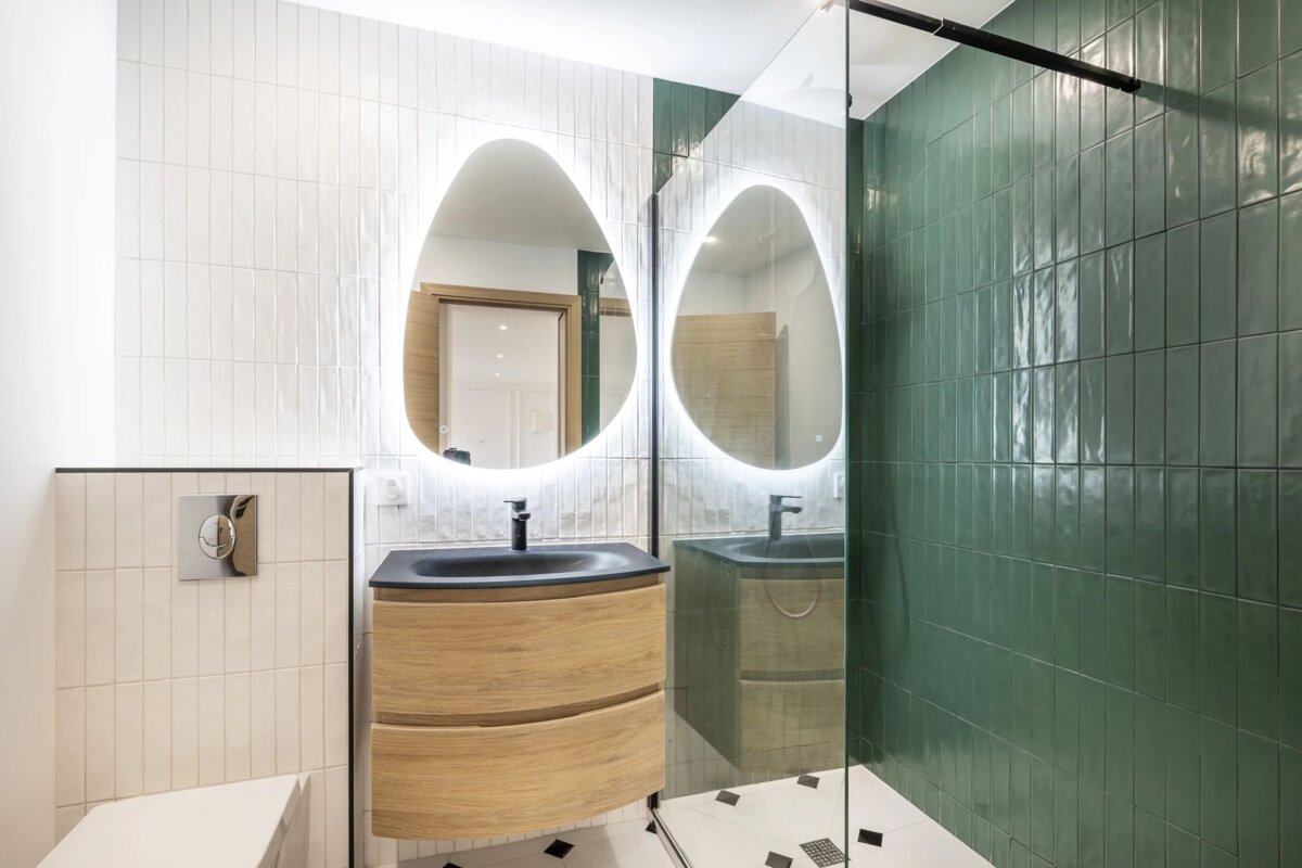 A modern bathroom with a wooden vanity, an illuminated pebble-shaped mirror, white wall tiles, and a green-tiled glass shower.