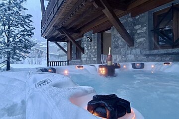 A steaming outdoor hot tub with champagne on a snowy winter day next to a rustic wooden chalet.