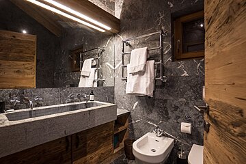 Rustic-modern bathroom featuring dark marble walls, a large stone trough sink, wooden accents, and a heated towel rack.