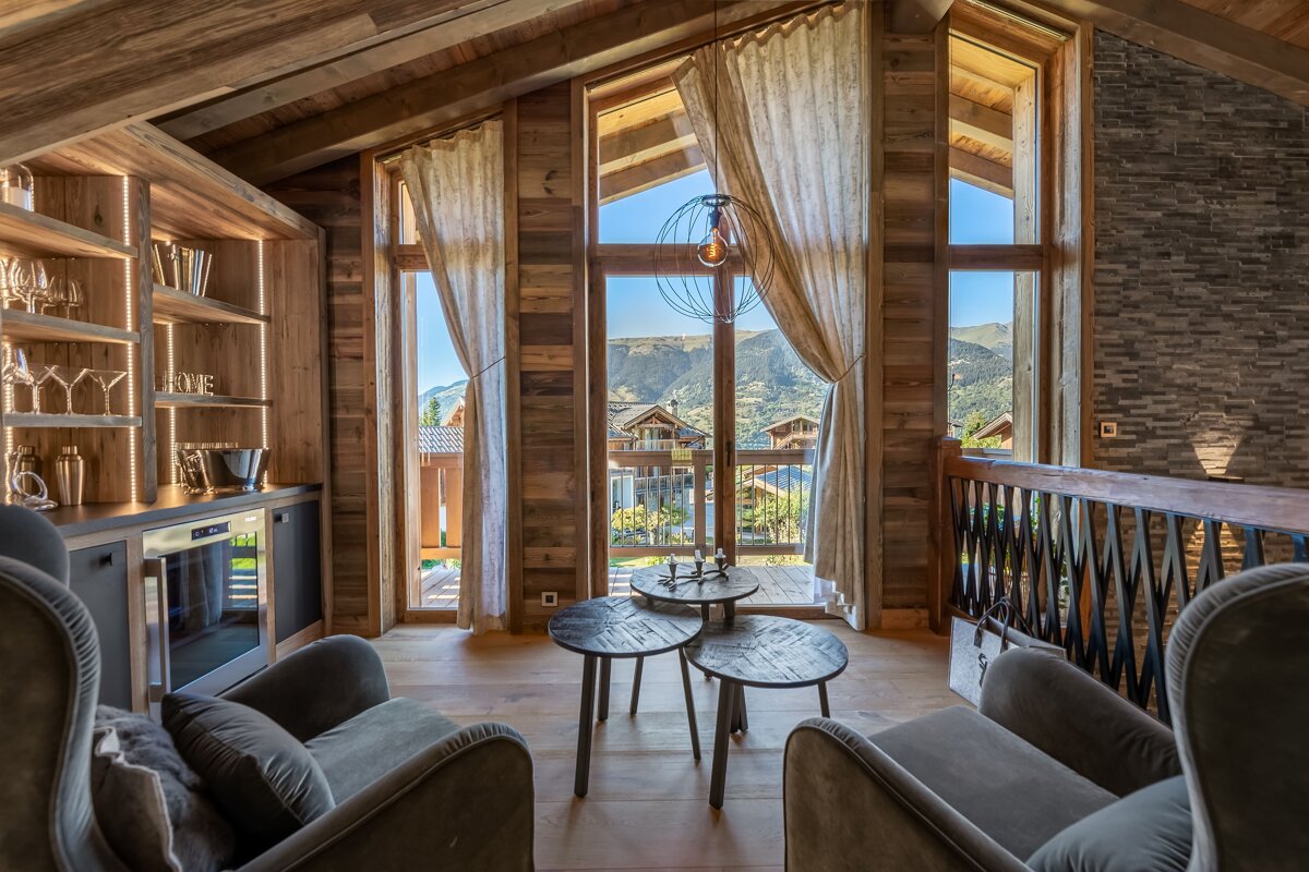 A luxurious, wood-paneled chalet interior with a bar, cozy seating, and large windows offering a stunning mountain village view.
