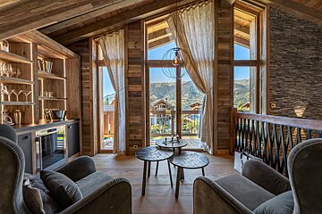 Cozy chalet interior with rustic wood walls, armchairs, nested tables, a bar, and large windows overlooking mountain views.
