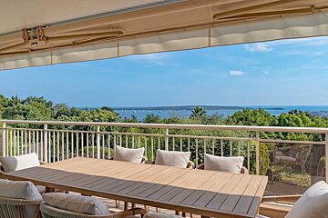 A spacious terrace with an outdoor dining set and awning, overlooking a beautiful bay dotted with boats and lush green trees under a blue sky.
