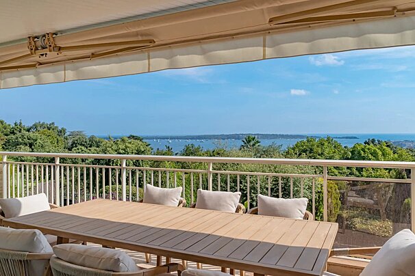 A wooden dining set on a balcony under an awning, overlooking lush greenery, the sea, and distant land under a blue sky.