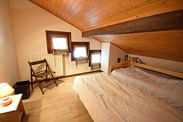 An attic bedroom featuring two beds, a sloped wooden ceiling with beams, several small windows with blinds, and light wood flooring.