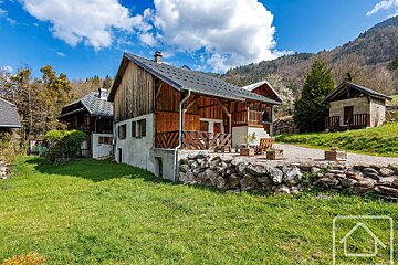 Alpine chalets with wooden details stand on a grassy slope, framed by mountains and a blue, cloudy sky.