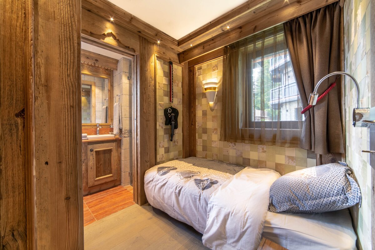 A cozy, wood-paneled bedroom featuring a single bed, window, warm lighting, and an en-suite bathroom entrance. Rustic chalet-style decor.