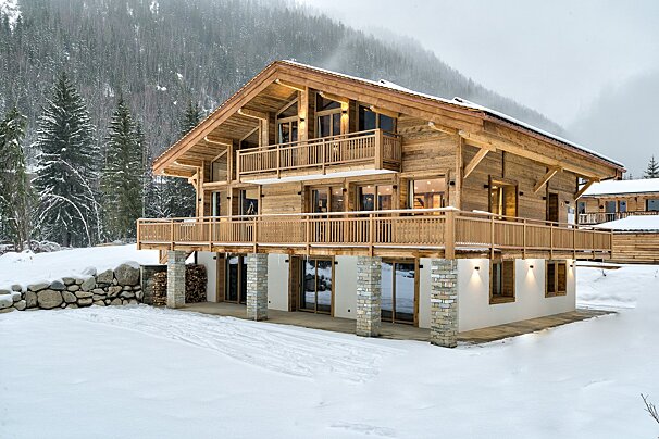 A grand, multi-story wooden chalet with balconies and large windows stands in a pristine, snow-covered mountain landscape with a dense pine forest.