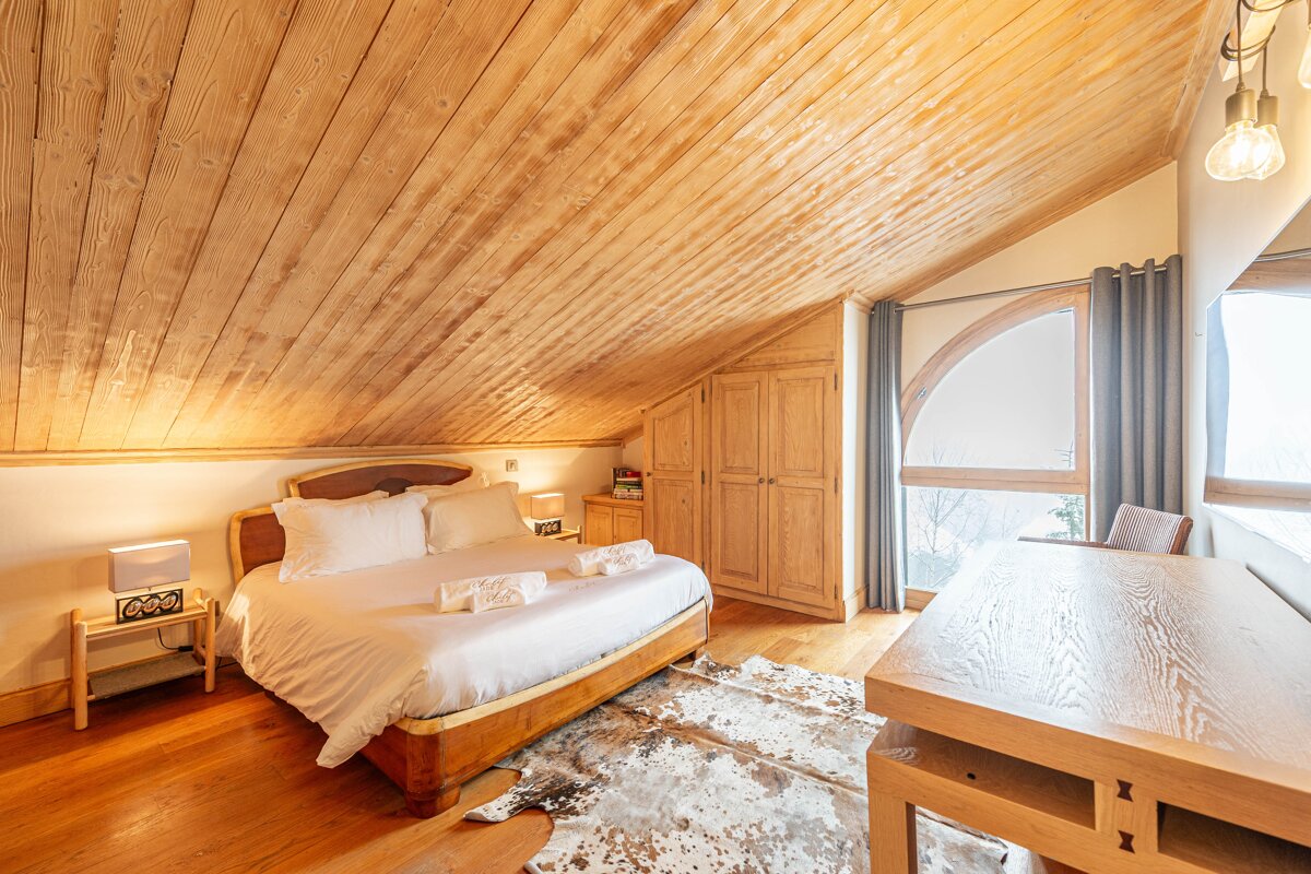 Cozy cabin-style attic bedroom featuring a sloped wood ceiling, large bed, cowhide rug, and an arched window.