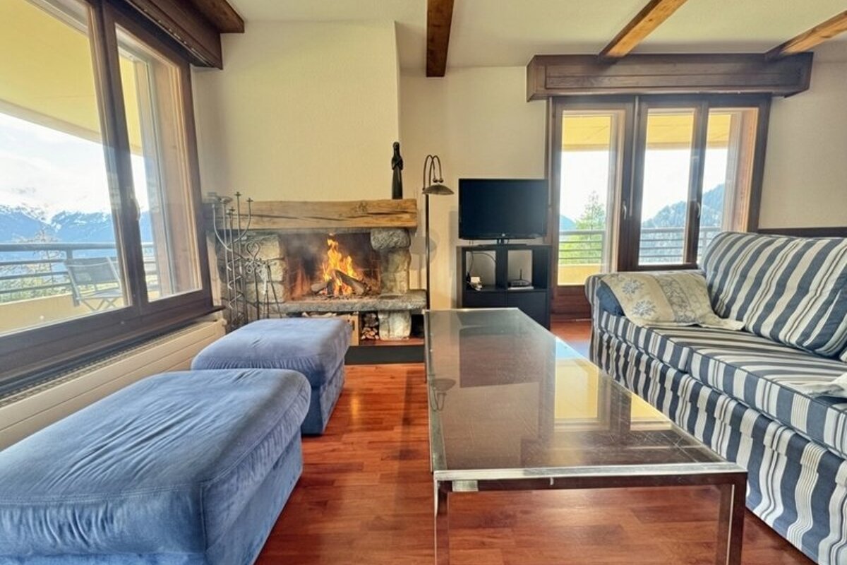 Cozy living room with a burning fireplace, striped sofa, blue ottomans, a glass coffee table, and scenic mountain views through large windows.