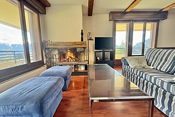 Cozy living room with a burning fireplace, striped sofa, blue ottomans, a glass coffee table, and scenic mountain views through large windows.