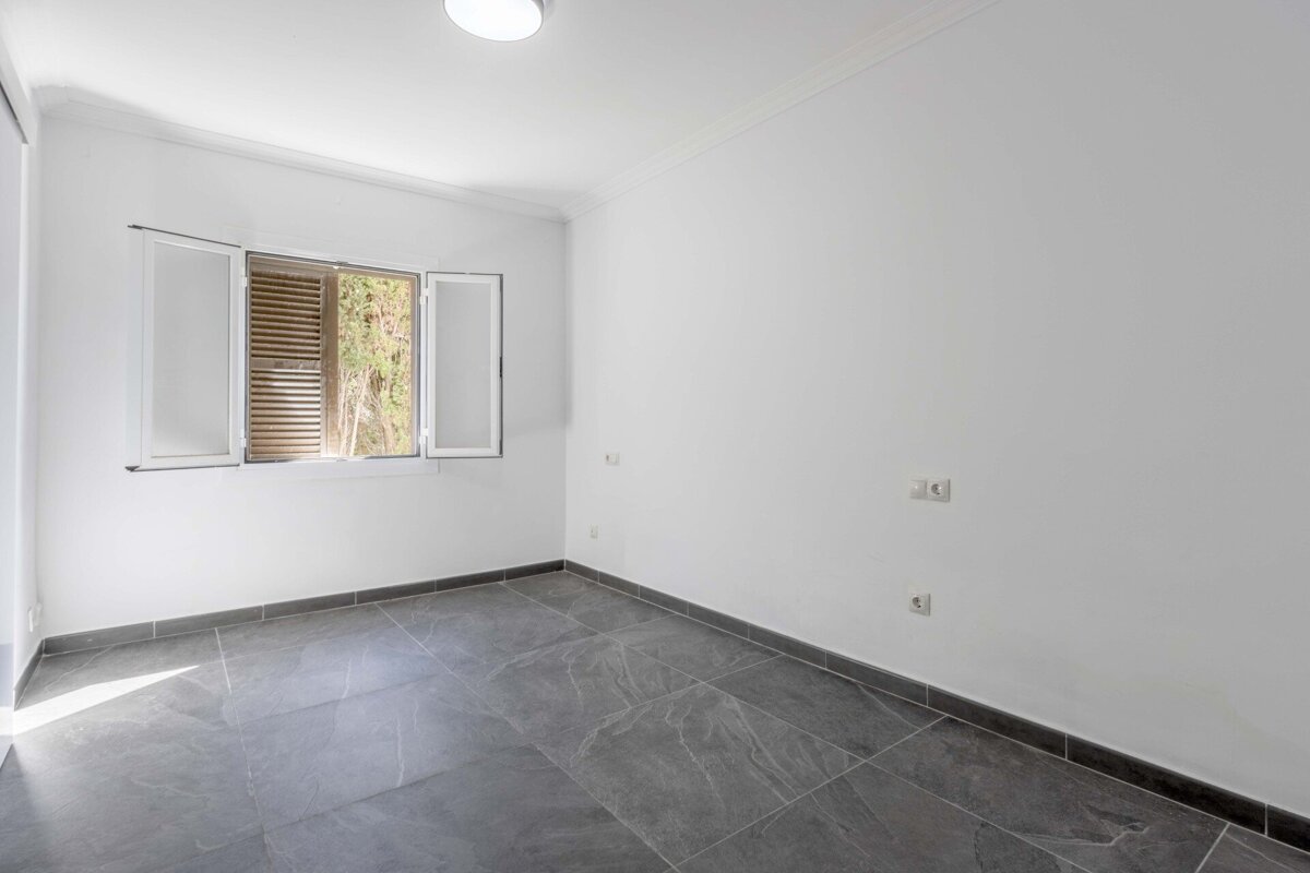 A bright, empty room with white walls, dark gray tiled floor, and a window with open shutters revealing greenery. A ceiling light and outlets are visible.