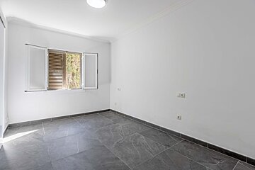 A bright, empty room with white walls, dark gray tiled floor, and a window with open shutters revealing greenery. A ceiling light and outlets are visible.