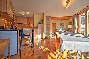 Rustic cabin interior featuring a kitchen with wooden cabinets, terracotta floor tiles, and a dining table bathed in warm sunlight.