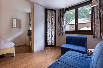 A room with wooden flooring features blue convertible seating, a large window revealing mountains and buildings, and patterned curtains. A separate area contains a single bed, TV, and mirror.