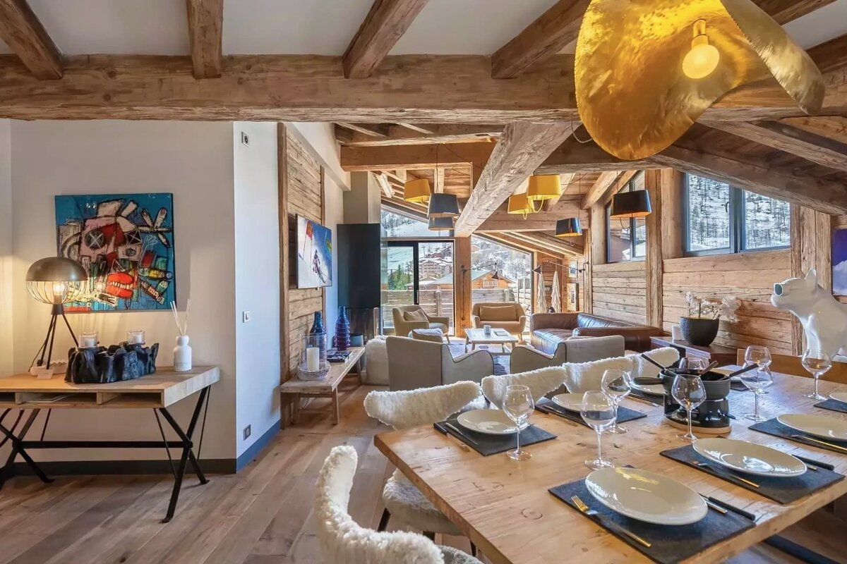 A luxurious open-plan chalet living and dining area with rustic wooden beams, modern art, and a snowy mountain view. A set dining table and cozy seating fill the space.