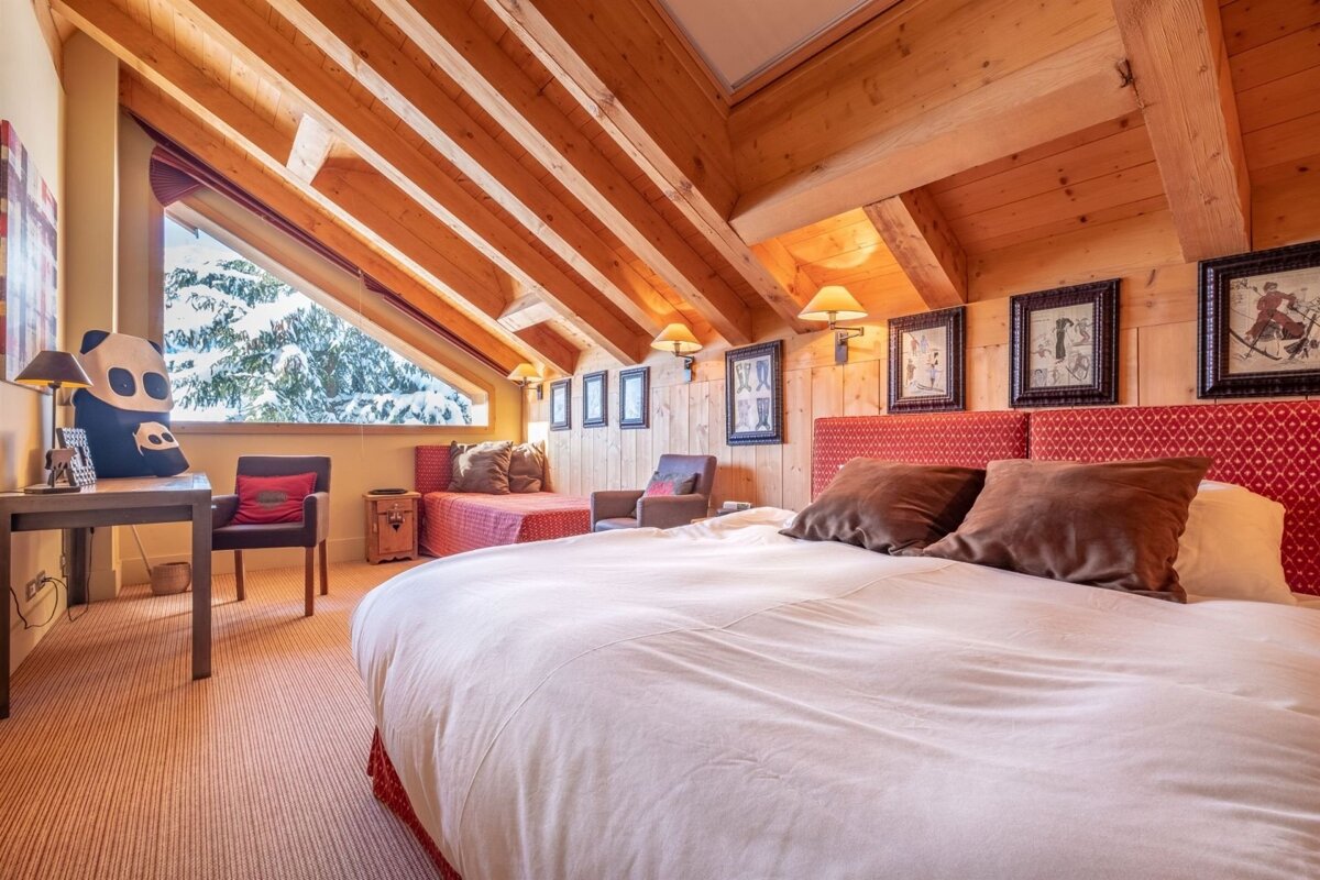 A cozy ski chalet bedroom features exposed wooden beams, a large bed with red accents, and a window overlooking snowy trees.
