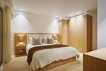 A modern, serene bedroom with a large bed, light wood finishes, neutral bedding, built-in wardrobe, and soft, recessed lighting.
