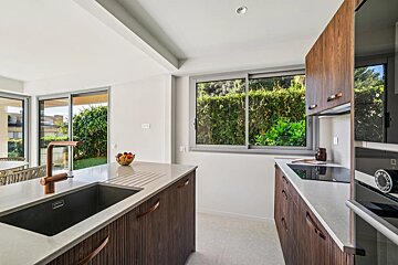 A modern kitchen with dark wood cabinets, light counters, and a copper faucet. Large windows and sliding doors offer views of lush green foliage.