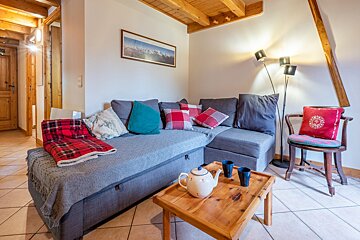 A warm and inviting living space featuring a grey sectional sofa with vibrant cushions, a wooden coffee table, and exposed timber ceiling beams.