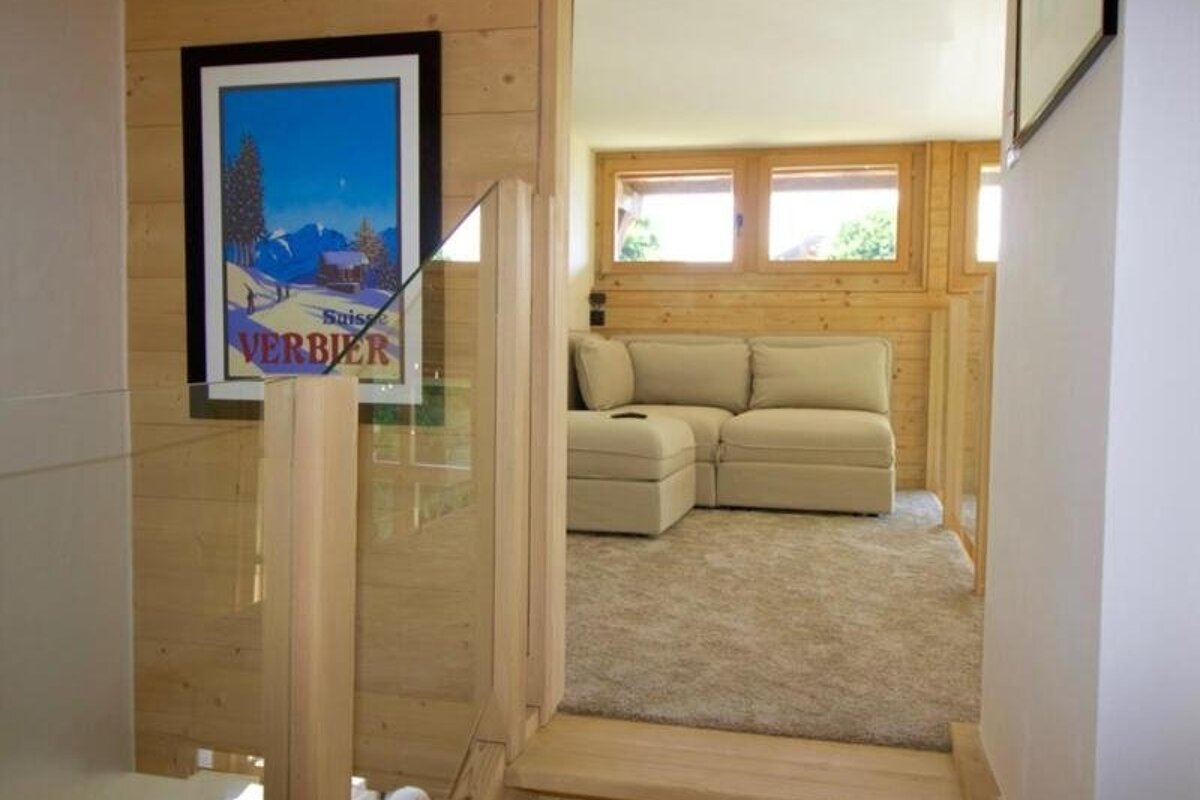 Cozy wood-paneled room with a beige sectional sofa, carpet, and a framed Verbier travel poster by a glass stair railing.