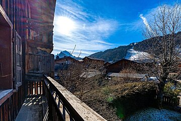 Sunny view from a rustic wooden balcony of a mountain village, chalets, and ski slopes under a bright blue sky.