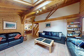 A large living room with abundant exposed wood beams & paneling, black leather sofas, tiled floor, and a dining area beyond.