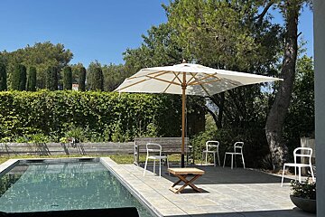 Sunny patio with a swimming pool, white umbrella, chairs, and lush green hedges under a clear blue sky.