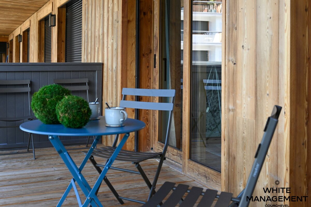 A cozy wooden balcony with a blue bistro table, mugs, and green decorative balls. Folding chairs and sliding glass doors complete the scene.
