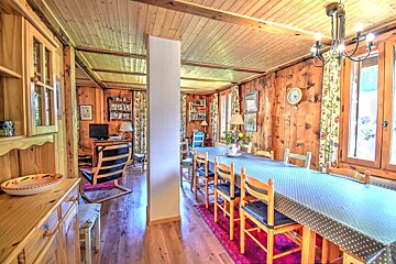 Rustic, wood-paneled dining room with a long table and chairs. A cozy living area with a bookshelf and TV is visible beyond a white pillar.
