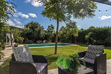 A vibrant, sunny backyard features a sparkling blue pool, lush green lawn, and comfortable patio seating. A white gazebo adds charm.