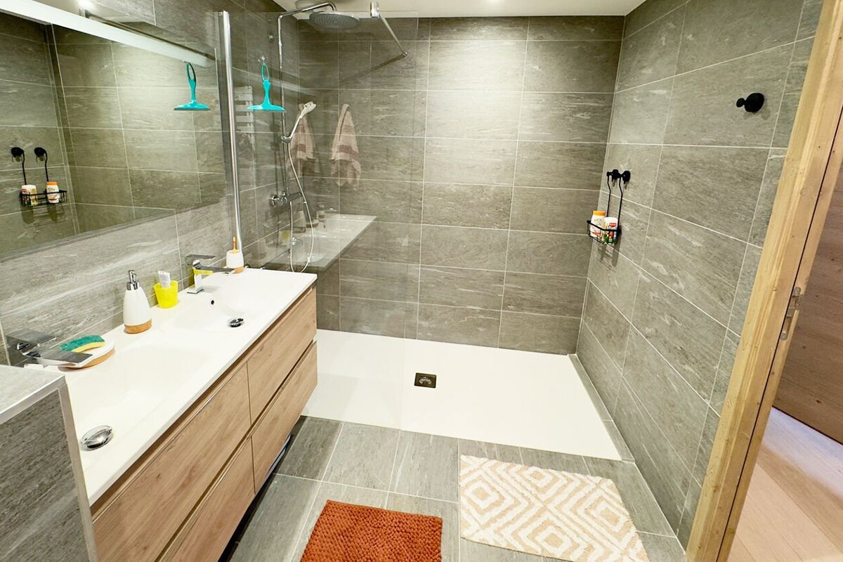 A bright, modern bathroom featuring gray tiled walls and floor, a double vanity, and a spacious walk-in shower with clear glass. Two bath mats add color.