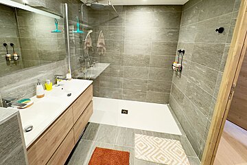 A bright, modern bathroom featuring gray tiled walls and floor, a double vanity, and a spacious walk-in shower with clear glass. Two bath mats add color.