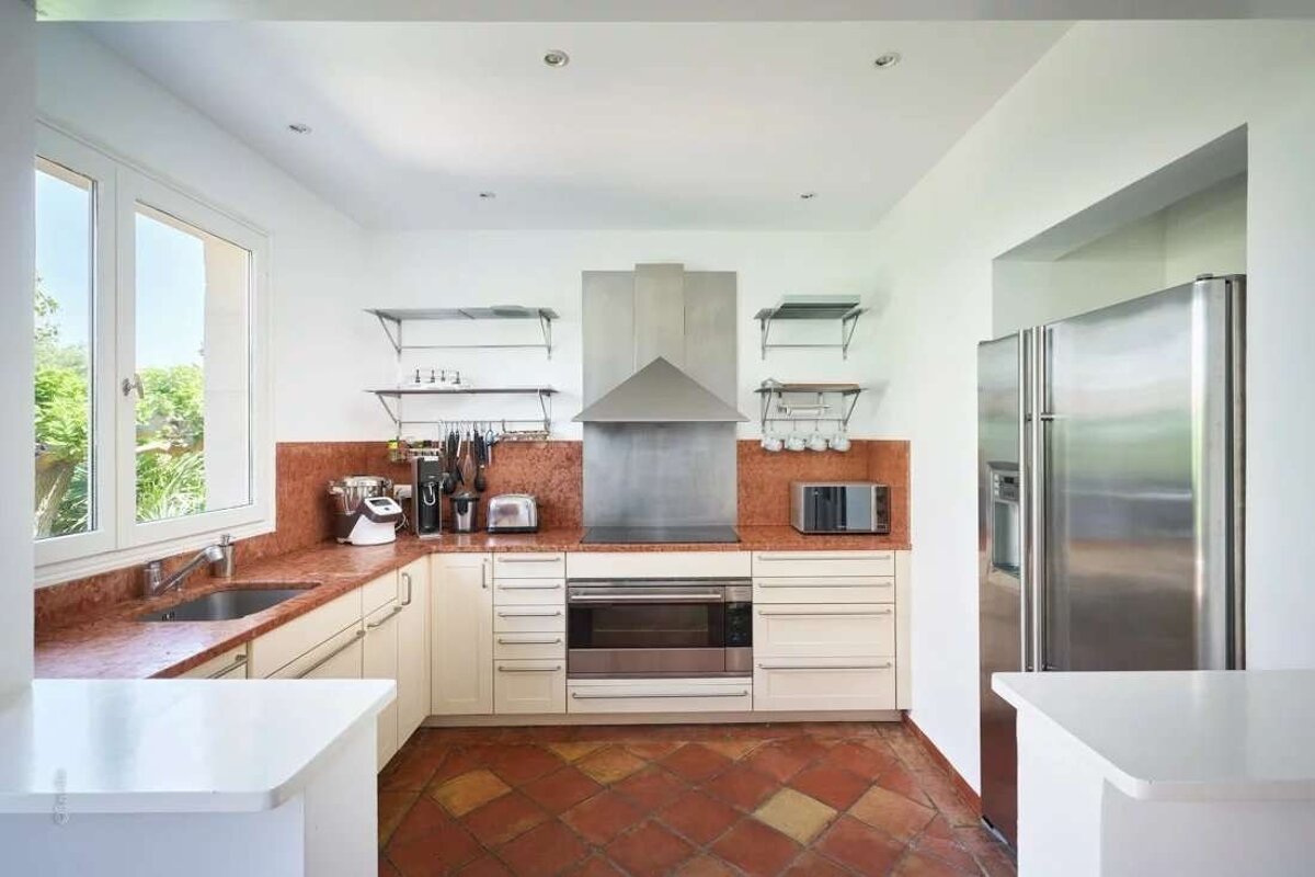 A bright, modern kitchen featuring red-toned tiled floors, cream cabinets, red marble countertops, and stainless steel appliances.