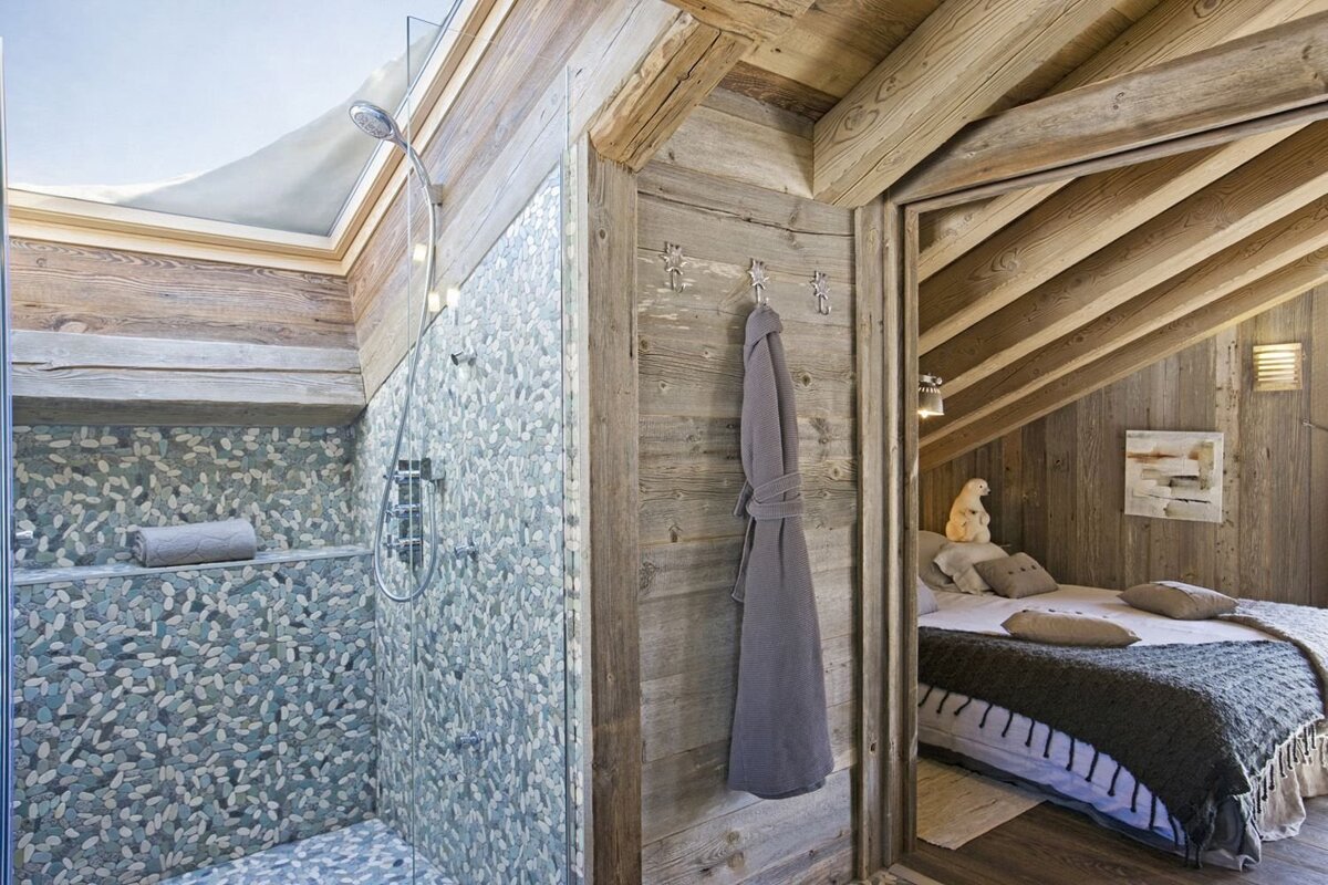 Cozy chalet interior with a pebble-tiled shower and glass screen adjacent to a rustic, wood-paneled bedroom under an angled ceiling.