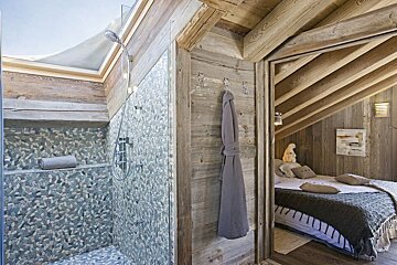 Cozy chalet interior with a pebble-tiled shower and glass screen adjacent to a rustic, wood-paneled bedroom under an angled ceiling.