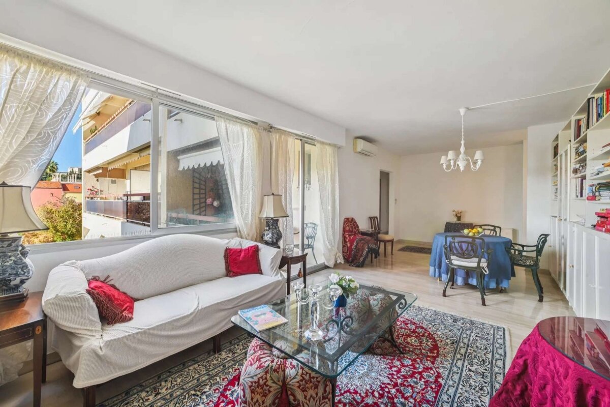 A bright, traditionally furnished living room with a white sofa, ornate rugs, a glass coffee table, and a dining area. Large windows overlook a balcony.