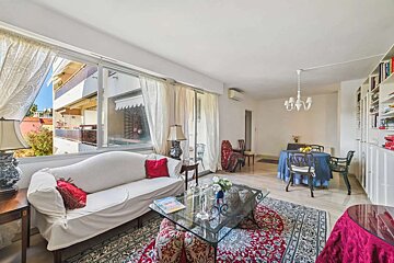 A bright, traditionally furnished living room with a white sofa, ornate rugs, a glass coffee table, and a dining area. Large windows overlook a balcony.