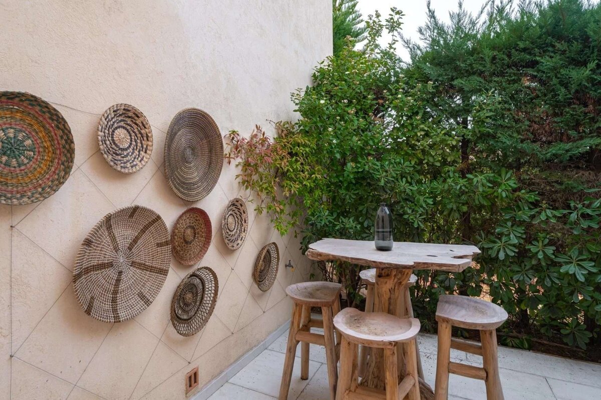 A rustic outdoor patio with decorative woven baskets on a wall, a wooden table and stools, surrounded by lush green foliage.