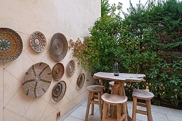 A rustic outdoor patio with decorative woven baskets on a wall, a wooden table and stools, surrounded by lush green foliage.