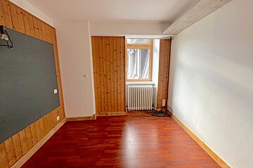 An empty room with dark wood flooring, white and wood-paneled walls, a window with a radiator, and a large dark board on one wall.