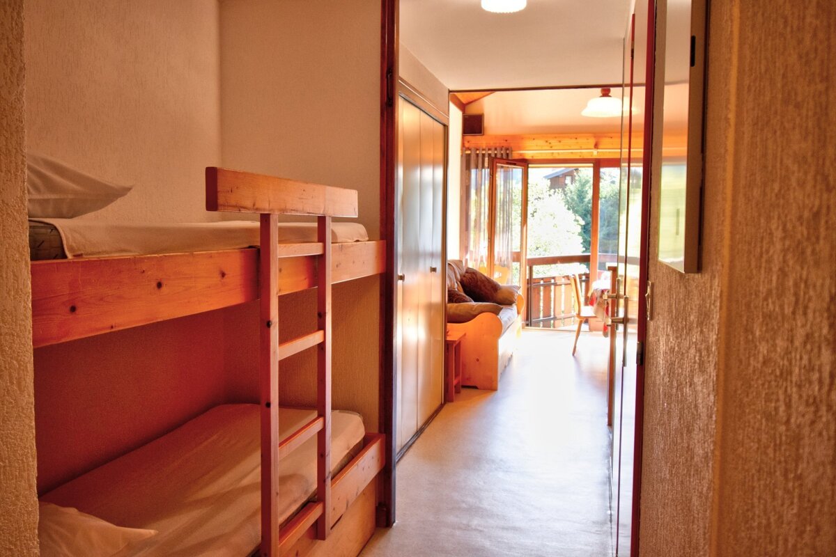 A room with wooden bunk beds on the left, opening into a bright living area with a sofa and a balcony offering an outdoor view.