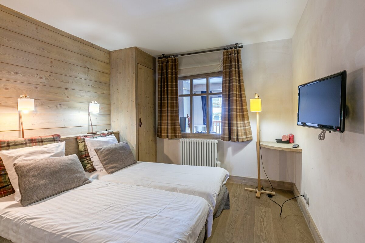 A rustic bedroom with twin beds, wood-paneled walls, plaid curtains, a window, a wall-mounted TV, and a yellow floor lamp.