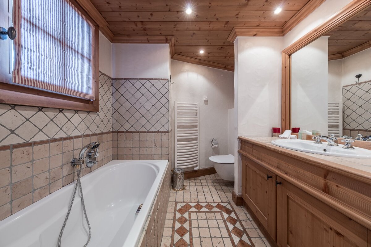 Rustic bathroom with a wooden ceiling, diamond-patterned tiled walls, decorative floor, bathtub, wooden vanity with sink, and toilet.
